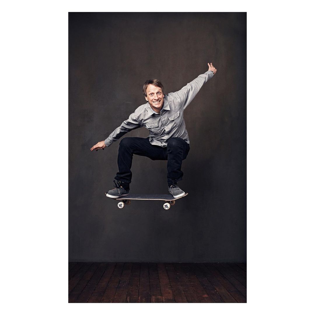 Adult male skateboarder, gray long sleeve shirt and dark jeans, mid-air jump, dark background, indoors, medium shot, skateboard in motion.