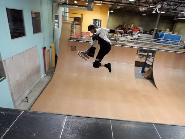 A skateboarder is airborne in a skatepark, wearing protective gear, riding a skateboard, and doing a trick in a wooden halfpipe.