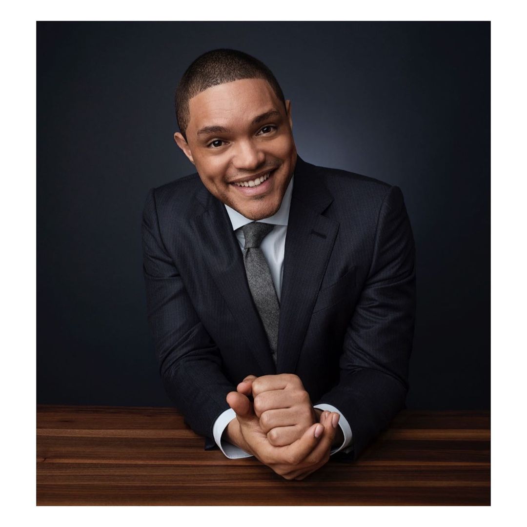 Portrait of a smiling man in a suit and tie sitting at a wooden table against a dark backdrop, looking directly at the camera.