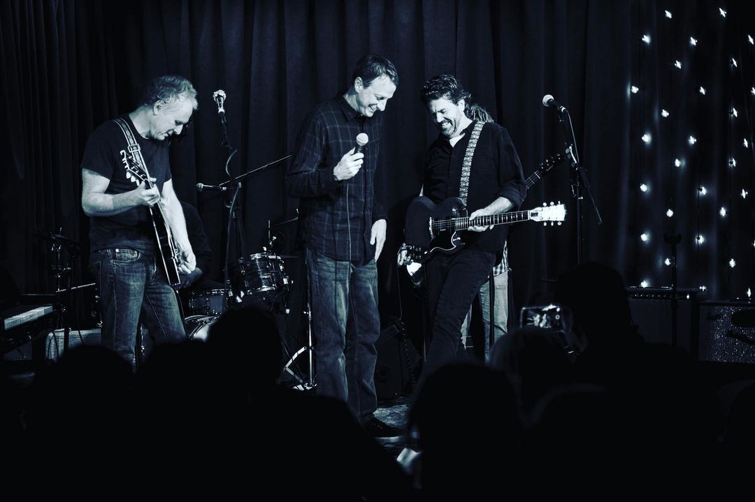 Three adult male musicians perform live on a stage with instruments and microphones, with a shadowy audience in the foreground.
