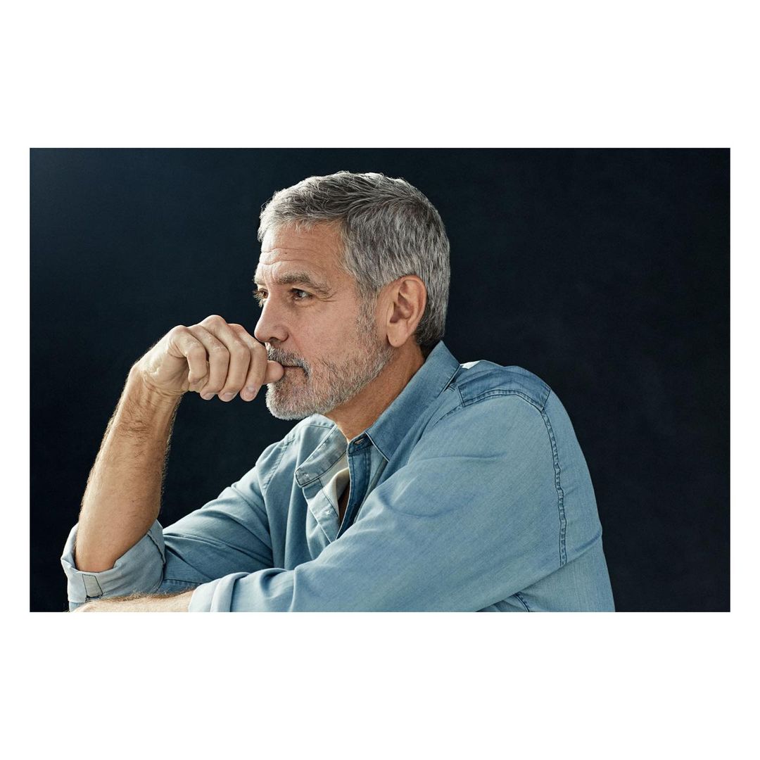 Mature adult man gazes thoughtfully in a profile shot. The man has graying hair and a light beard, wearing a blue denim shirt, against a dark backdrop.
