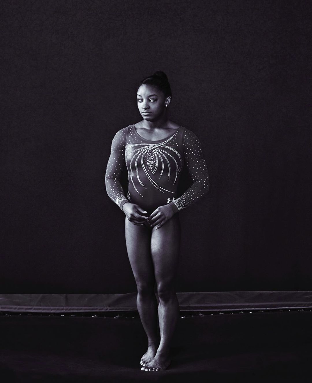 Full-length portrait of Simone Biles in a leotard with long sleeves, posed against a dark background on a trampoline.