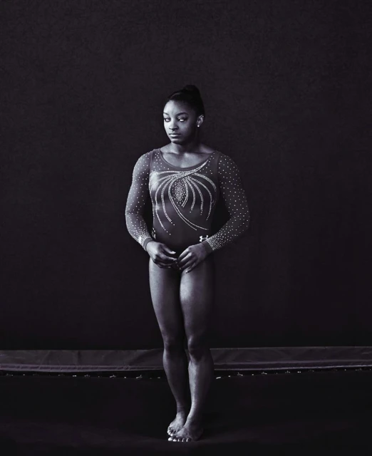 Full-length portrait of Simone Biles in a leotard with long sleeves, posed against a dark background on a trampoline.