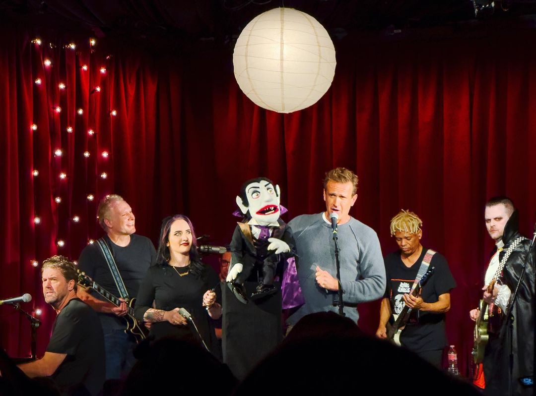 A group performs on a stage with a puppet, including musicians playing guitars, a singer, and a person dressed as a vampire.