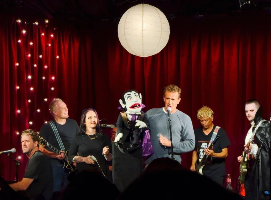 A group performs on a stage with a puppet, including musicians playing guitars, a singer, and a person dressed as a vampire.