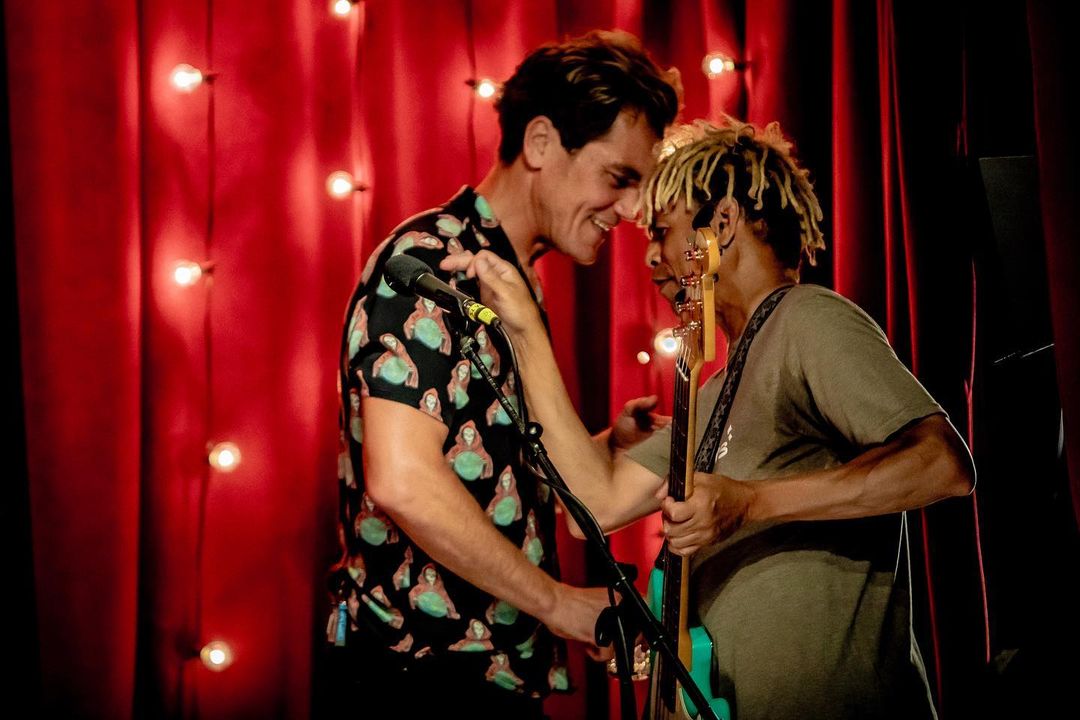 Two musicians face each other on stage in front of a red curtain and soft lighting. One holds a bass guitar and the other a microphone.