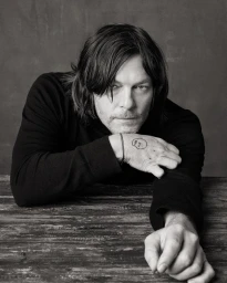 Portrait of Norman Reedus leaning on a wooden table wearing a black shirt against a textured dark background