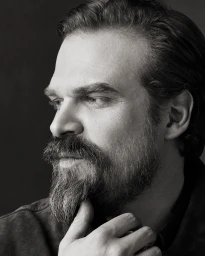 Black and white portrait of David Harbour with a full beard resting his chin on his hand.