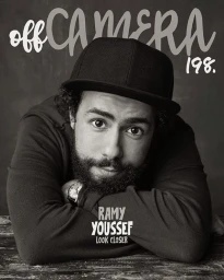 Black and white portrait of man with a beard wearing a baseball cap resting his arms on a textured surface, photographed by Sam Jones