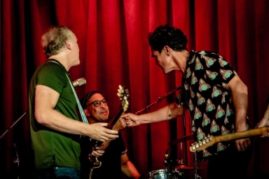 Three musicians on stage bumping fists in front of deep red velvet stage curtains, captured by photographer Sam Jones.