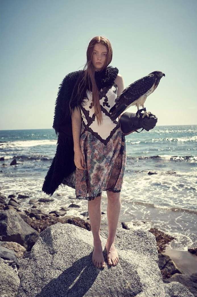 A young woman stands barefoot on a rock at the beach, holding a falcon on her gloved hand. She wears a patterned dress and dark cape.