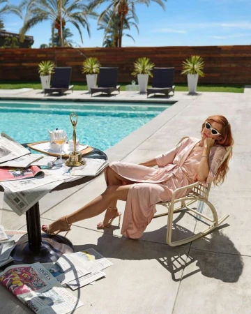Red-haired woman in pink silk robe lounging by a swimming pool in a white rocking chair with scattered newspapers