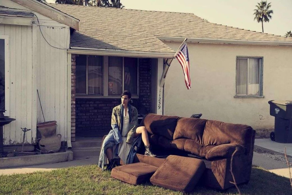 A man sits on an old couch placed on a lawn in front of a house with an American flag hanging from it. Palm trees are in the background.