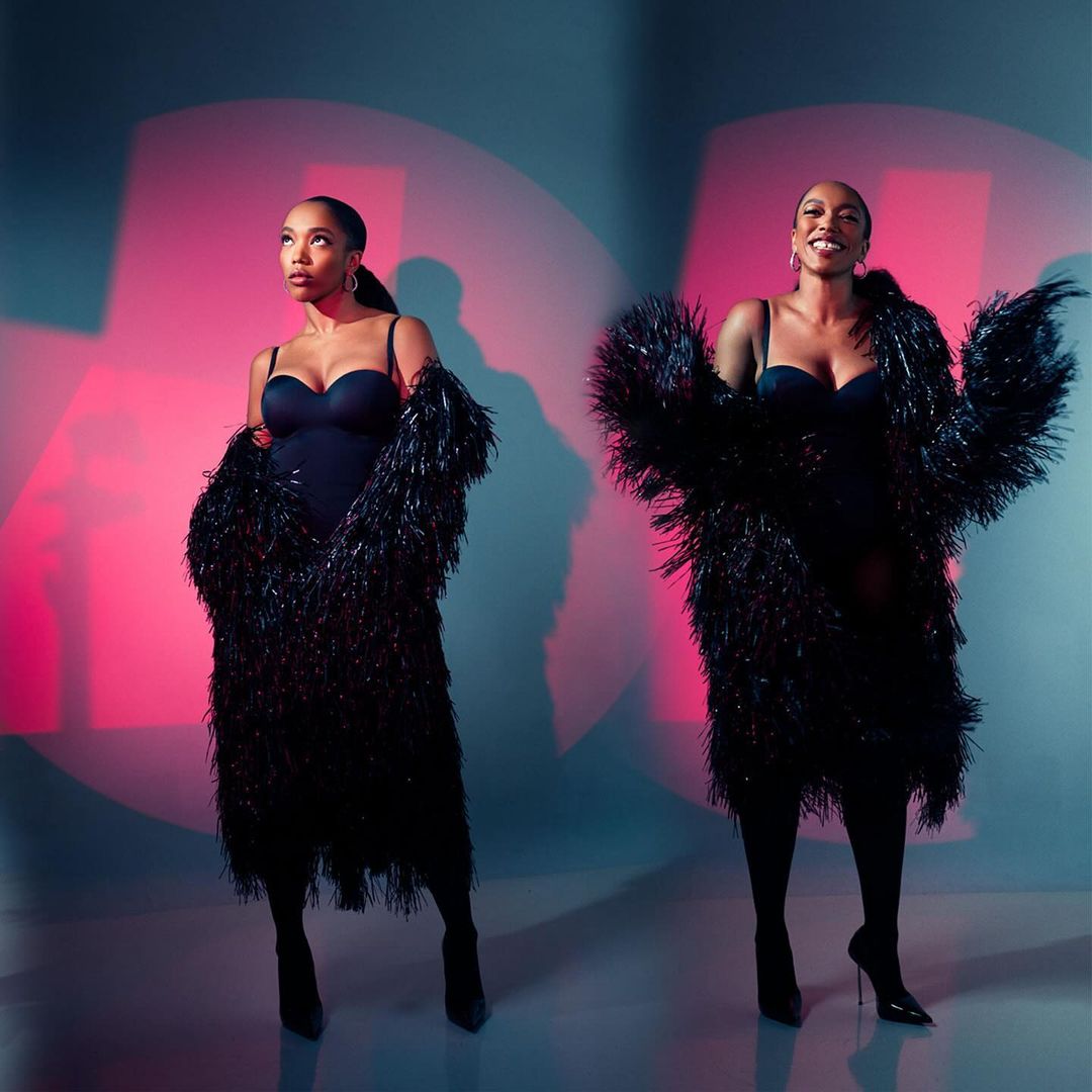 Two adult women pose in a photo studio wearing black bodysuits, textured black jackets, and heels against a pink and blue backdrop.