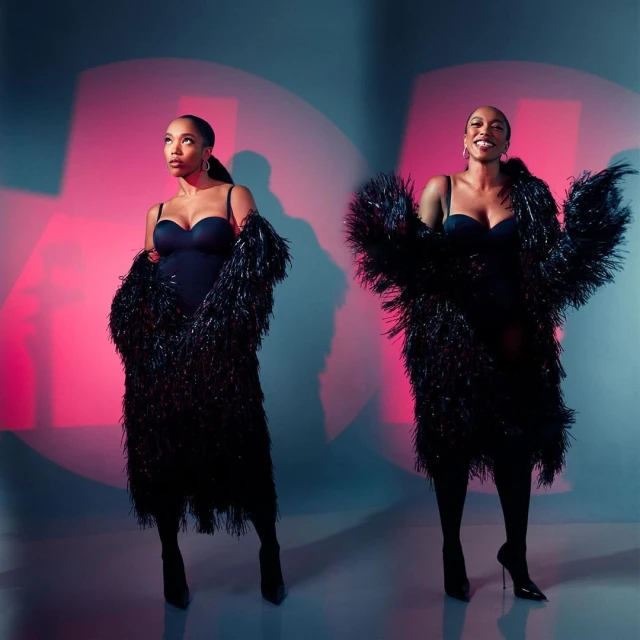 Two adult women pose in a photo studio wearing black bodysuits, textured black jackets, and heels against a pink and blue backdrop.