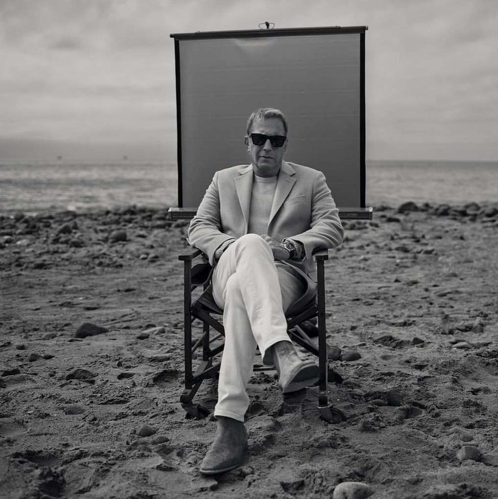 Adult male in suit seated in director's chair on beach with portable screen, neutral color palette, outdoor setting, and natural lighting.
