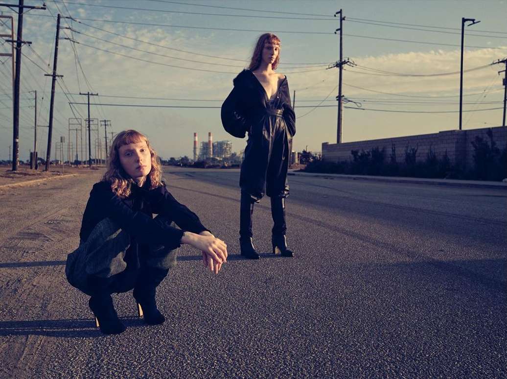 Two fashion models pose on an empty road. One model kneels, and the other stands, wearing dark, loose-fitting clothing. Background shows a cityscape.