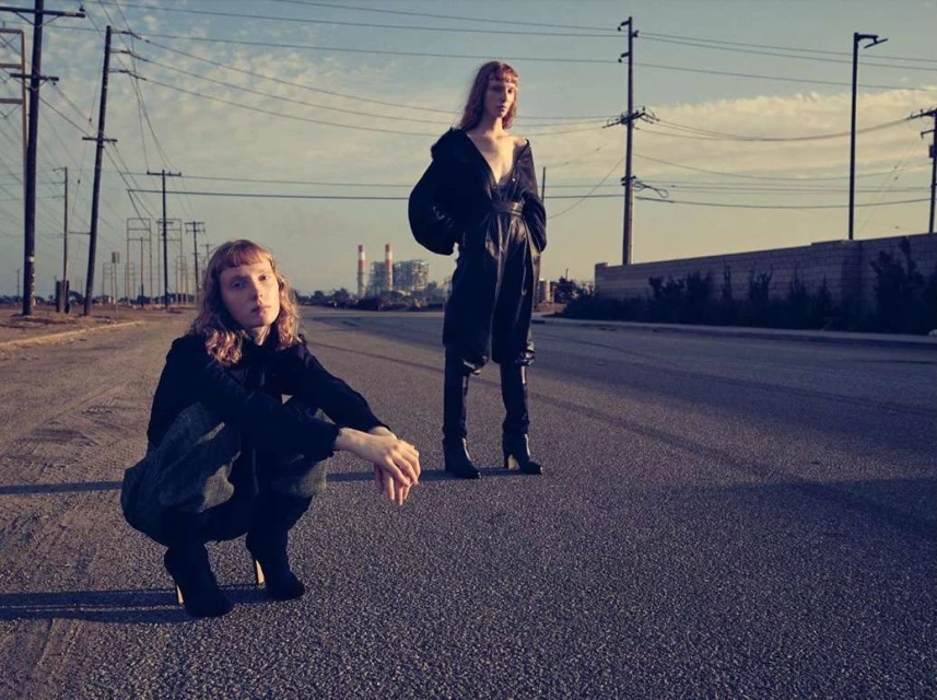 Two fashion models pose on an empty road. One model kneels, and the other stands, wearing dark, loose-fitting clothing. Background shows a cityscape.