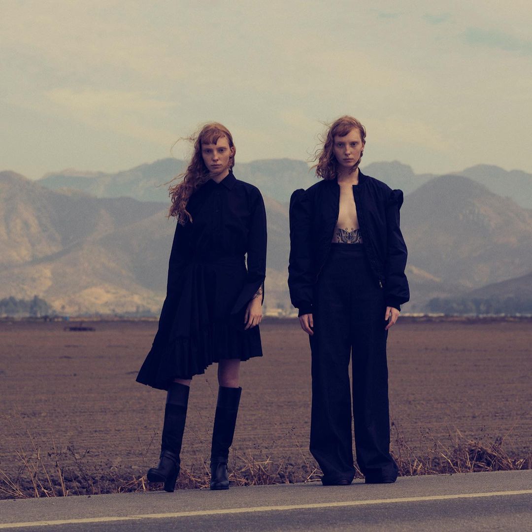 Two red-haired women stand in a field wearing black outfits, with a mountain range backdrop under a cloudy sky, creating a stark editorial image.