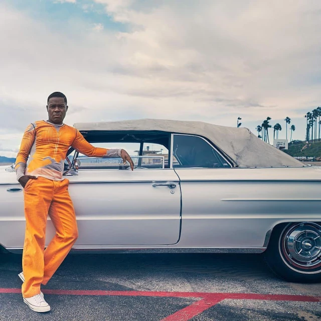 Adult male in orange outfit leaning against vintage car with palm trees and cloudy skies in the background, creating a casual fashion scene.