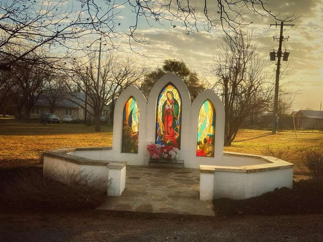 Outdoor view of a white memorial structure with three stained-glass windows depicting a woman, set in a grassy, rural area.