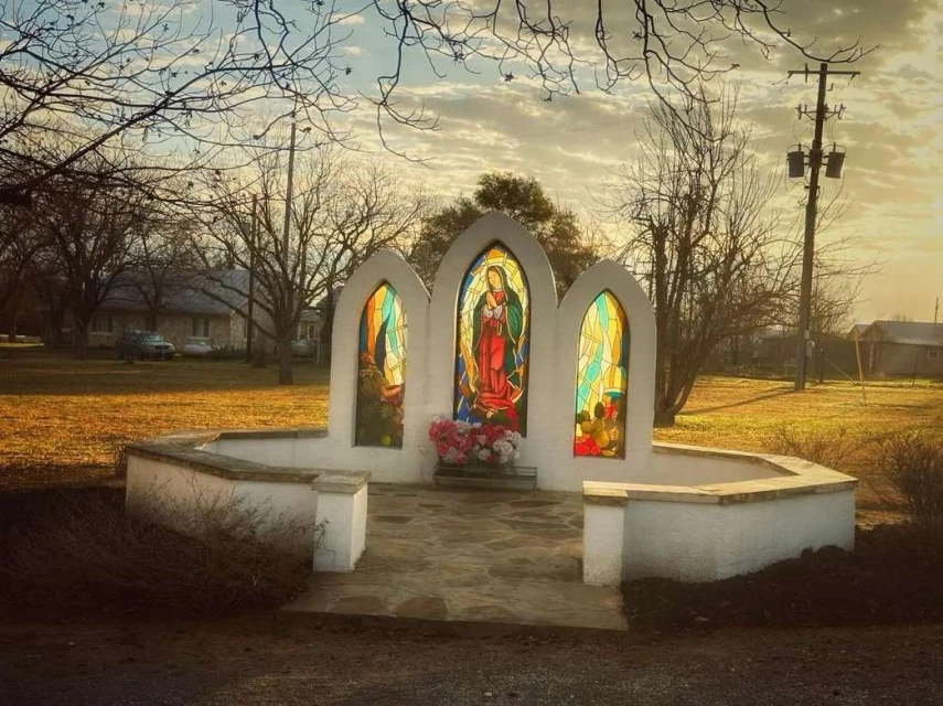 Outdoor view of a white memorial structure with three stained-glass windows depicting a woman, set in a grassy, rural area.