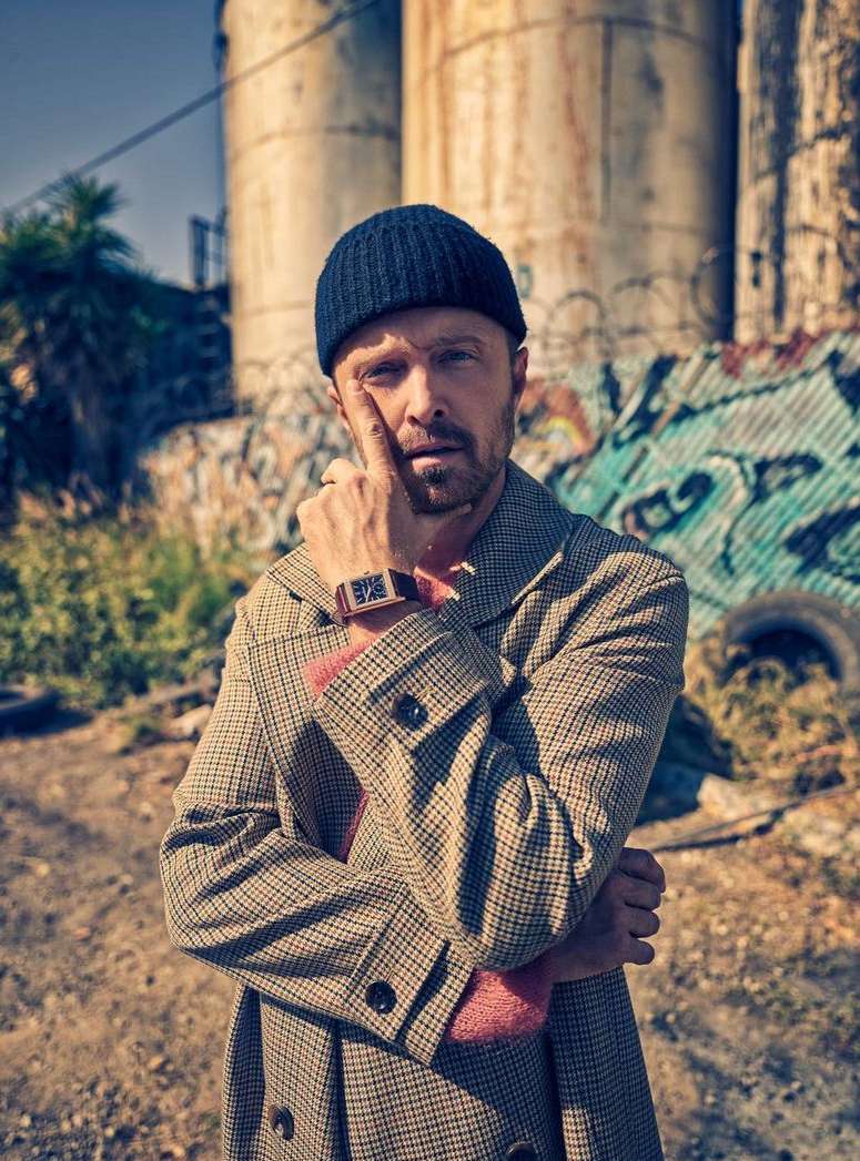 Portrait of a man wearing a beanie and patterned coat standing outdoors with silos and graffiti in the background on a sunny day.