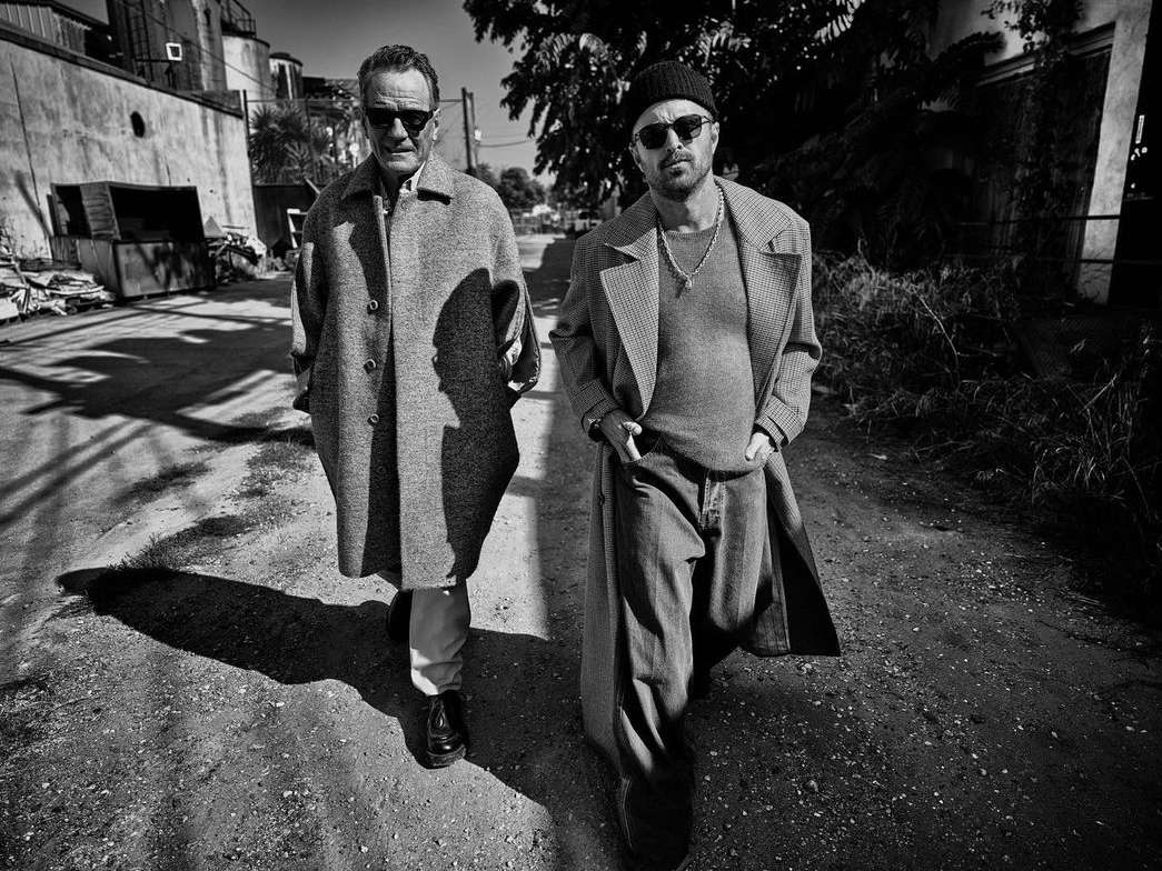 Two men in coats walk on a dirt road in a black and white photo. The taller man wears sunglasses, the other a beanie.