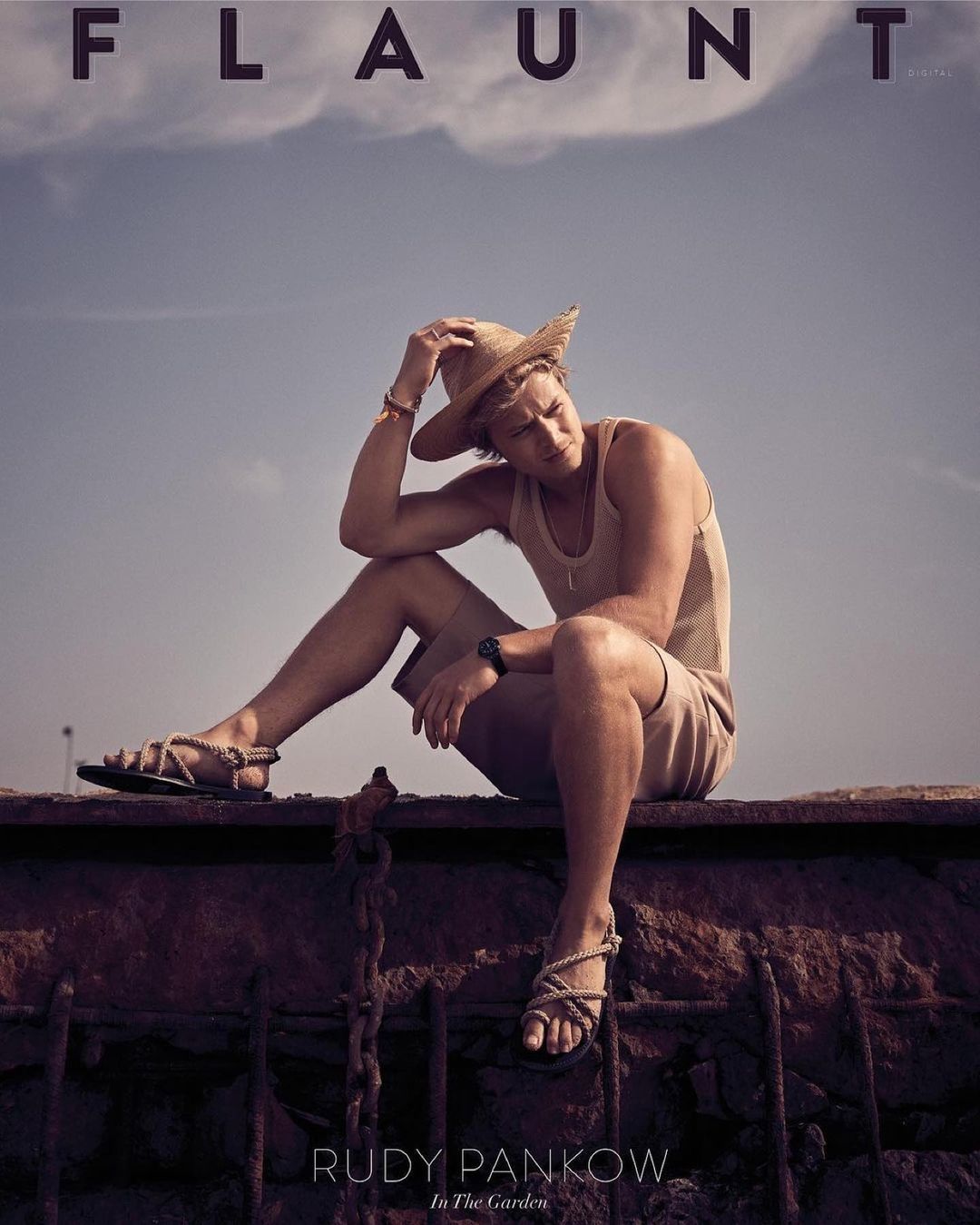 Rudy Pankow, model, poses outdoors wearing a tank top, shorts, sandals, and a straw hat in a casual fashion editorial shot.