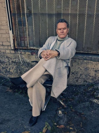Bryan Cranston seated outdoors in a light grey coat and turtleneck, hands clasped, against a wire mesh screen and concrete wall.