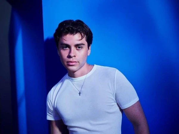 A young man wearing a white t-shirt and silver cross necklace posing against a vibrant blue wall.