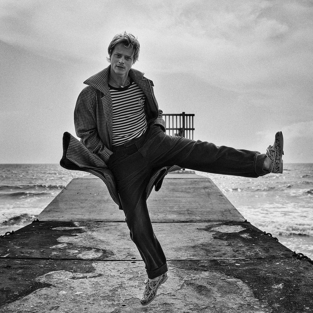 Black and white portrait of a man in a coat and striped shirt, mid-air on a concrete pier, with ocean and cloudy sky backdrop.