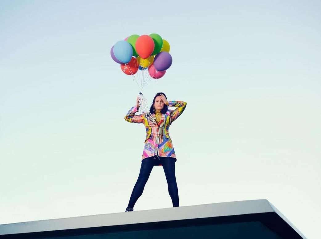 Woman in colourful patterned blazer standing on a rooftop holding a bunch of brightly coloured balloons against a clear sky.