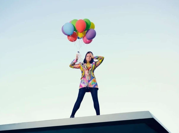Woman in colourful patterned blazer standing on a rooftop holding a bunch of brightly coloured balloons against a clear sky.