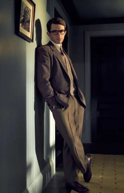 Portrait of a man in a brown suit leaning against a wall, wearing glasses, with an art piece visible in the background.