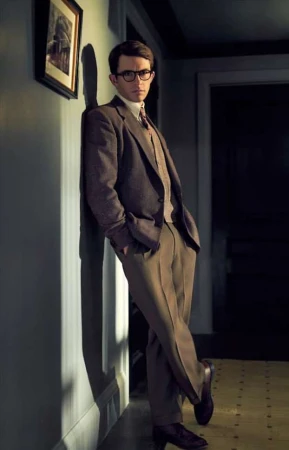 Portrait of a man in a brown suit leaning against a wall, wearing glasses, with an art piece visible in the background.