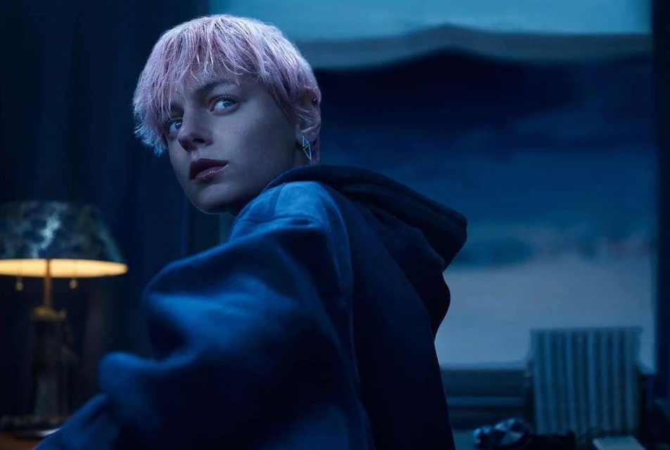 Portrait of a young adult with short, light pink hair looking over their shoulder, wearing a dark hoodie in a dimly lit room.