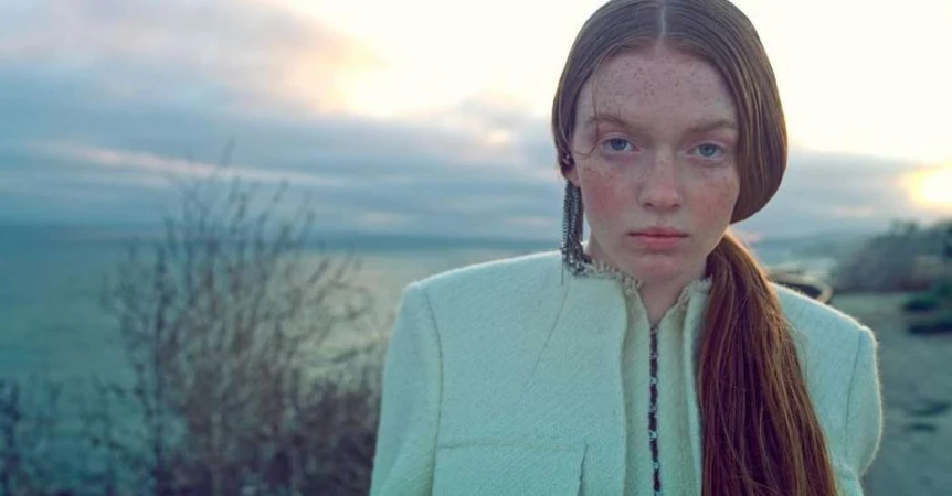 Portrait of a red-haired woman in a light green textured jacket against an overcast sky and coastal background.