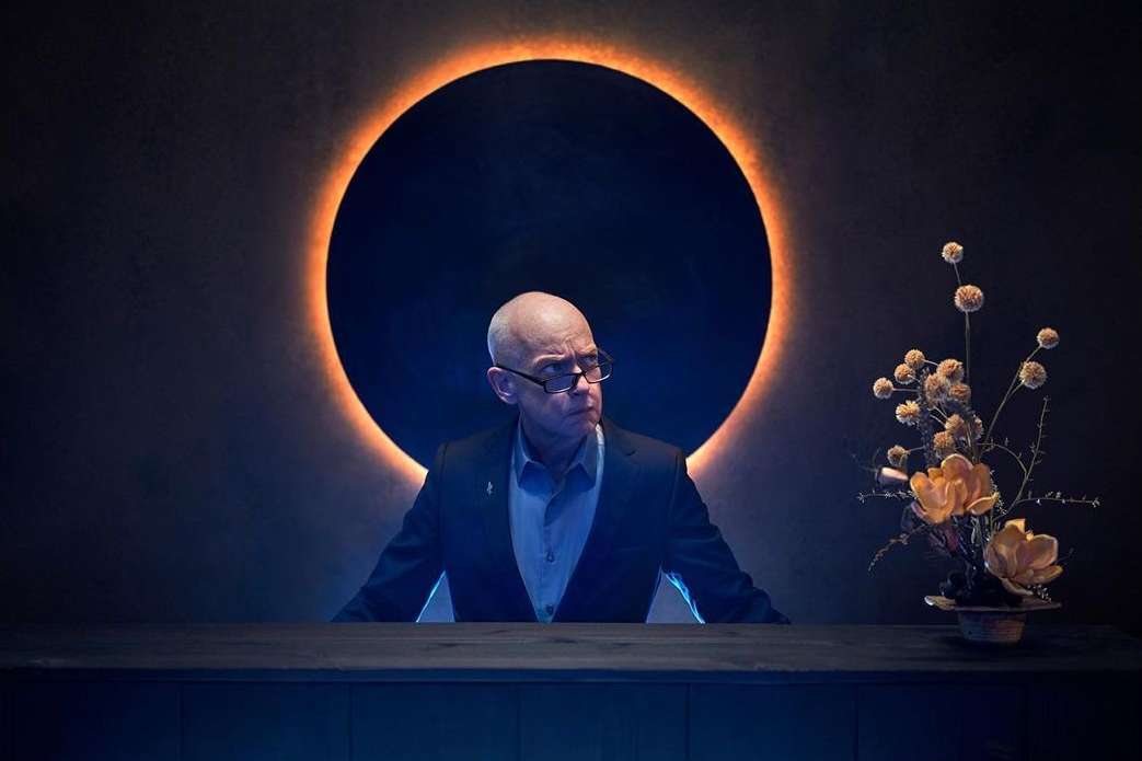 An adult male with glasses and a black suit sits at a dark table against a backdrop of a ring of light and a decorative floral arrangement.