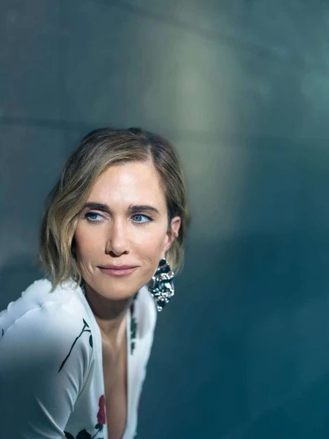 Close-up portrait of Kristen Wiig with short blonde hair, statement earrings, and a white floral-print dress against a textured gray background.