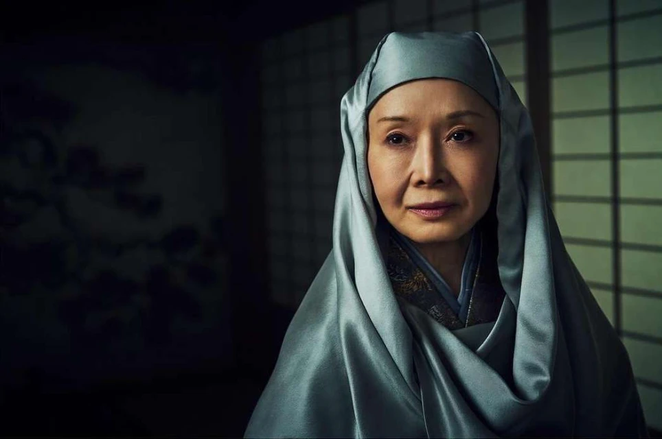 Close-up portrait of an adult female in a light blue head covering indoors. She is positioned in front of a dark background.