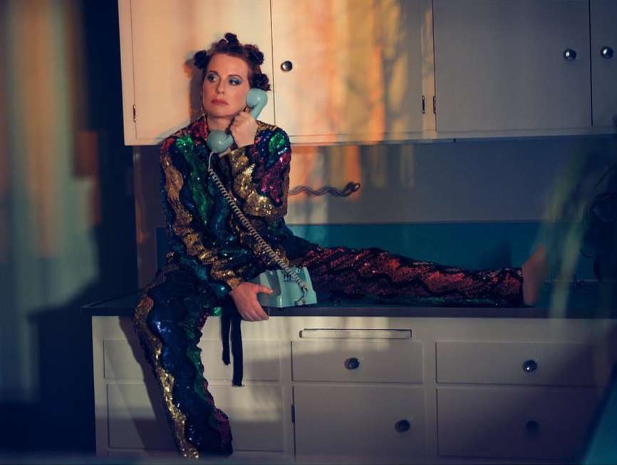 Woman wearing a sequined jumpsuit sits on a kitchen counter talking on a retro telephone in an indoor environment with colorful lighting.