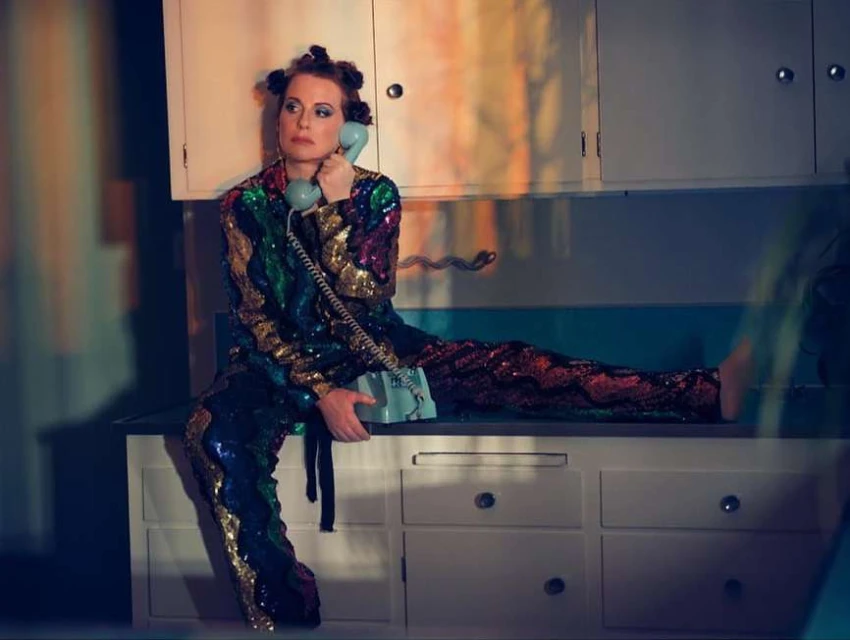 Woman wearing a sequined jumpsuit sits on a kitchen counter talking on a retro telephone in an indoor environment with colorful lighting.