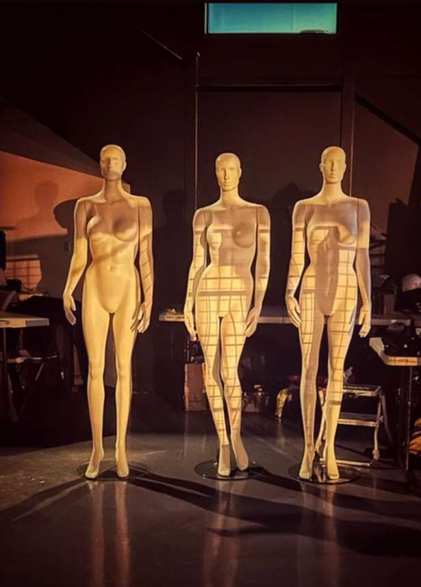 Three mannequins stand in a dark room, illuminated by a warm spotlight, creating long shadows on the floor. The mannequins appear to be in a studio.