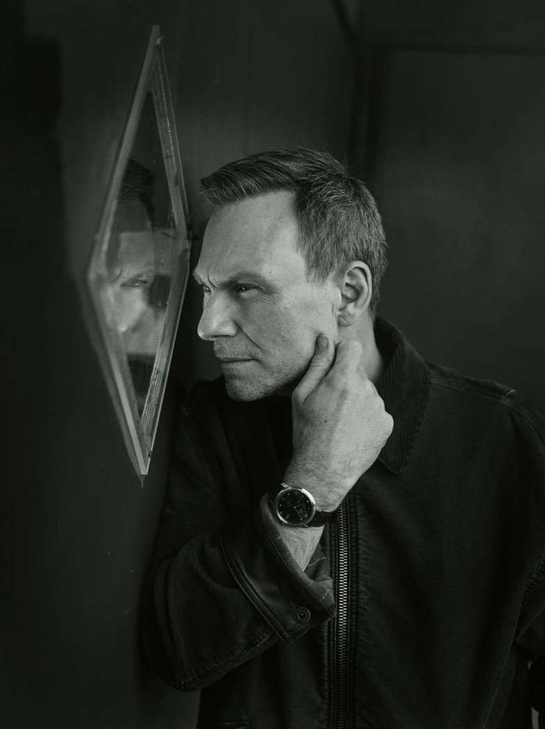 Monochrome portrait of a man touching his chin, looking intently into a diamond-shaped mirror on a dark wall