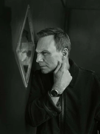 Monochrome portrait of a man touching his chin, looking intently into a diamond-shaped mirror on a dark wall