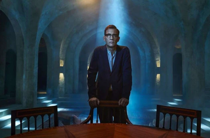 Man in glasses and a dark blazer standing behind a wooden chair inside an arched stone chamber lit by a blue overhead spotlight