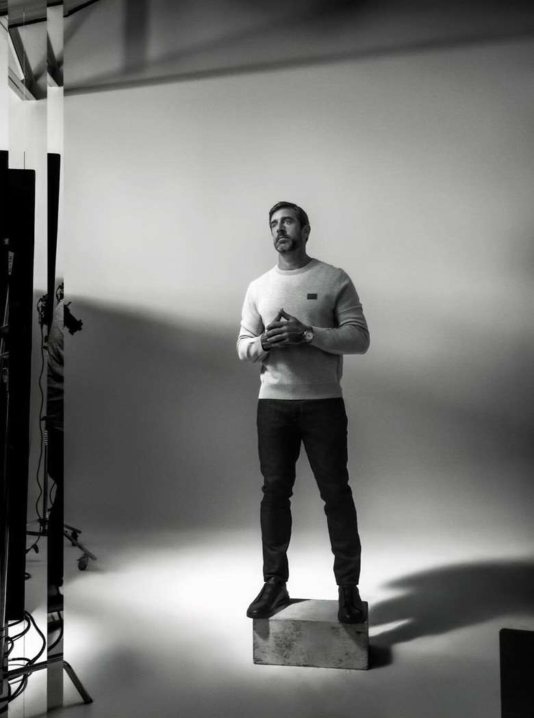 Monochrome full-body shot of a man in a sweater and jeans, standing on a box in a photo studio, looking upwards with his hands clasped.
