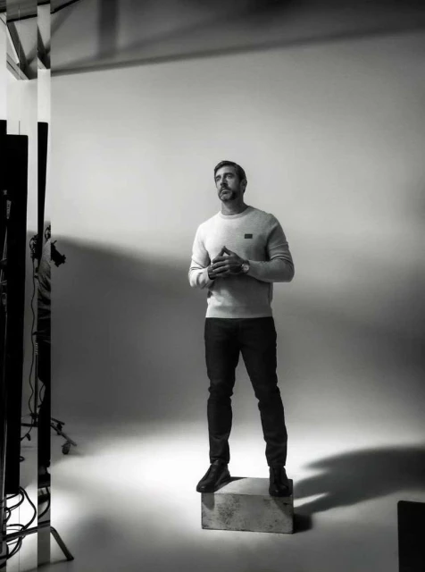 Monochrome full-body shot of a man in a sweater and jeans, standing on a box in a photo studio, looking upwards with his hands clasped.