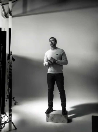 Monochrome full-body shot of a man in a sweater and jeans, standing on a box in a photo studio, looking upwards with his hands clasped.